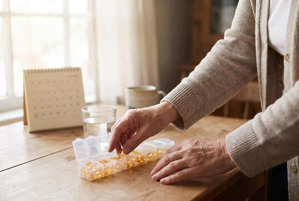 Older adult filling a weekly pill organizer with softgels at a wooden table beside a glass of water and a desk calendar