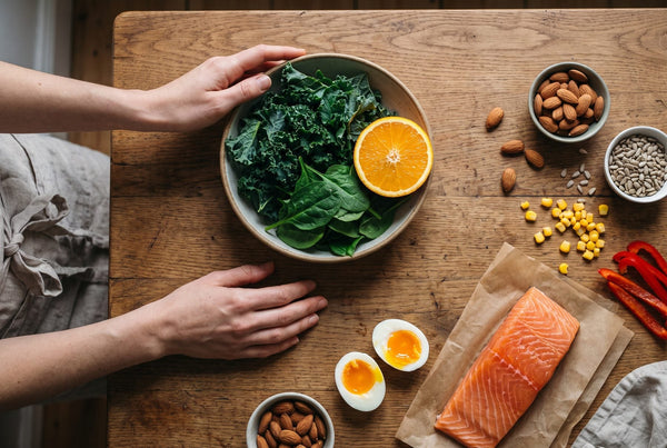 Wooden table with a bowl of kale, spinach and orange, plus salmon, eggs, almonds, sunflower seeds, corn and red pepper