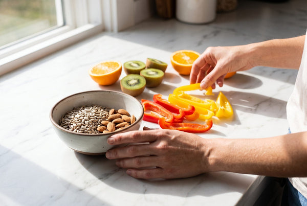 Hands arranging sliced red and yellow peppers, halved oranges and kiwifruit beside a bowl of almonds and sunflower seeds