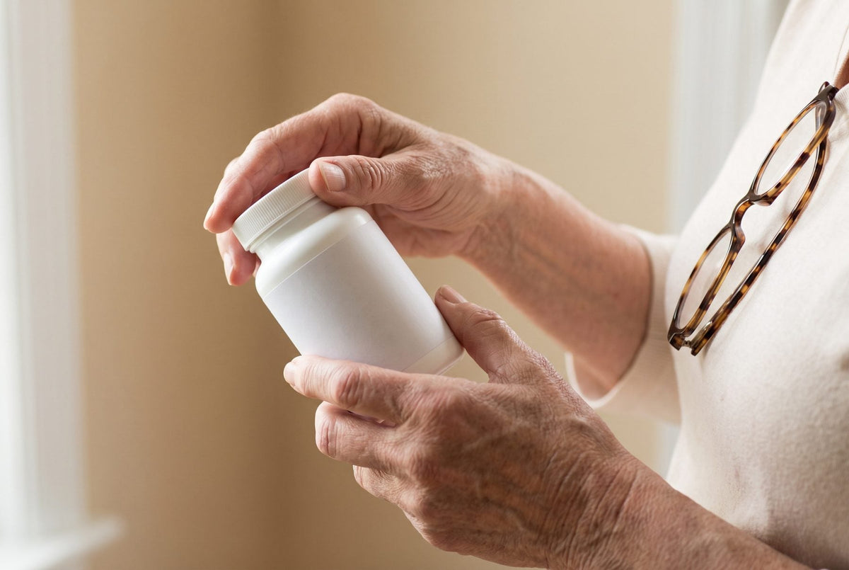Close-up of an older adult's hands holding a plain white supplement bottle, with reading glasses hanging from their collar