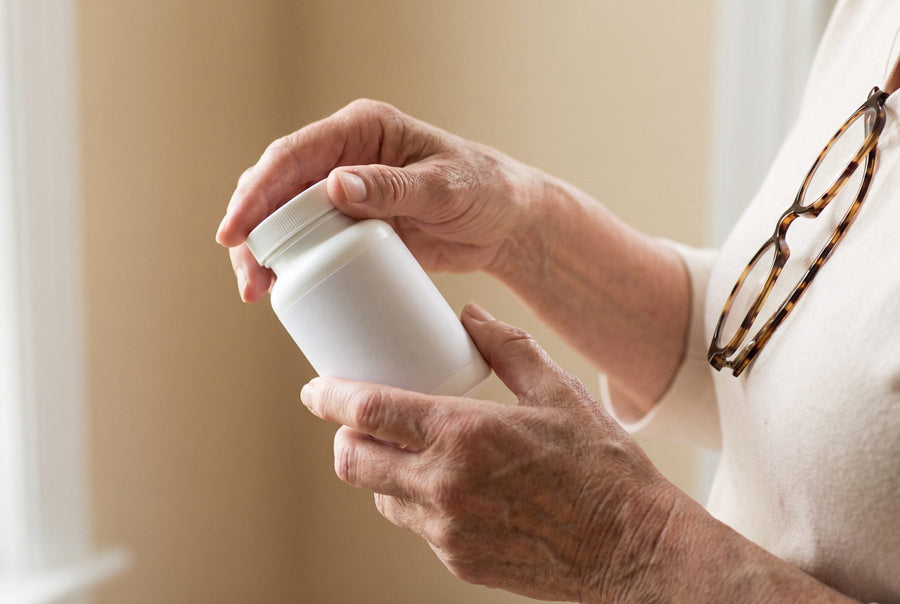 Close-up of an older adult's hands holding a plain white supplement bottle, with reading glasses hanging from their collar