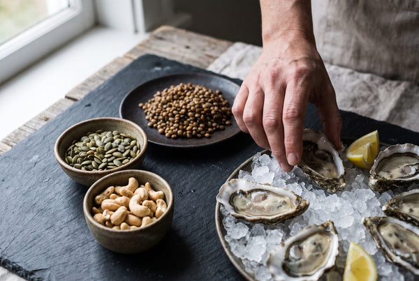 Fresh oysters on ice beside small bowls of pumpkin seeds, cashews and lentils on a slate board